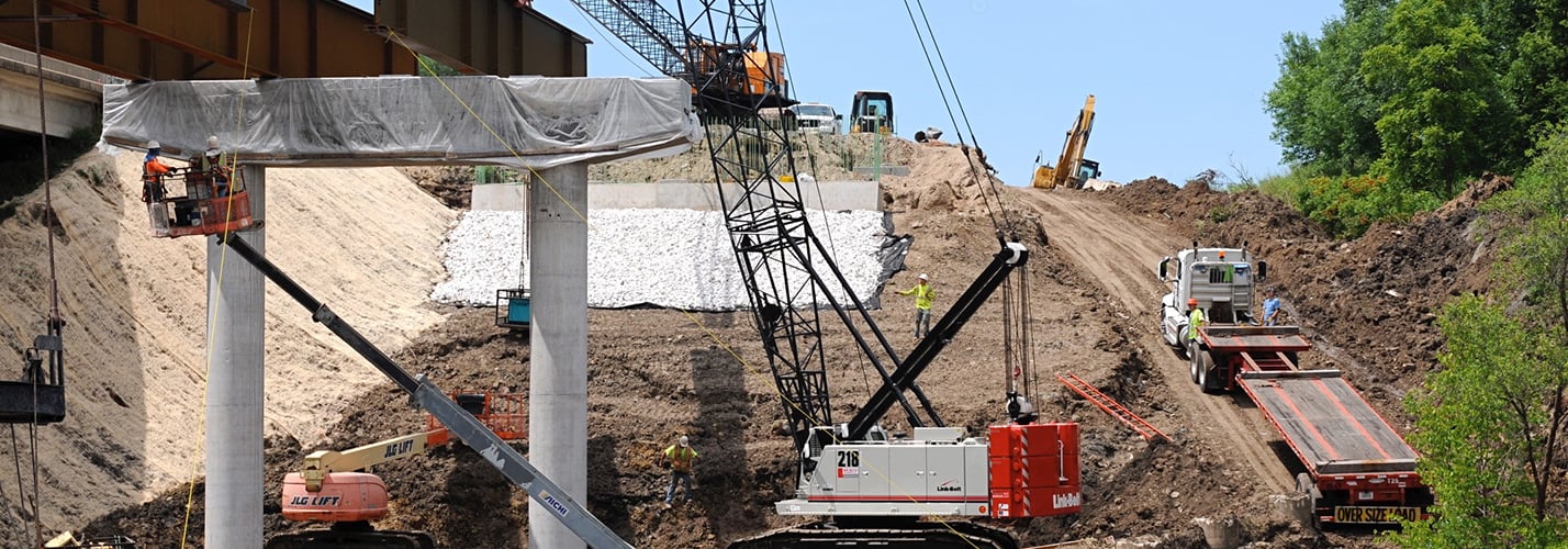 Bridge construction site with a crawler crane setting steel beams over concrete piers